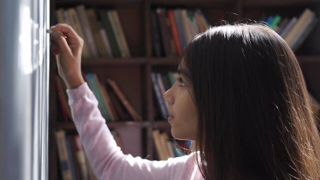 Serious indian kid primary school girl holding chalk writing on blackboard. Focused latin child preteen schoolgirl learning english alphabet letter handwriting standing in classroom. Close up view.