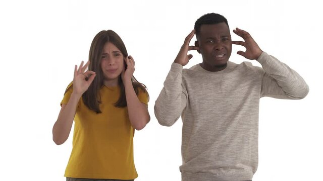 Young interracial couple suffering because of loud noise. African man and Caucasian woman plugging ears with fingers and begging to make sound quieter. Isolated on white background