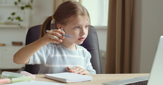 Portrait of little cute preschool girl wears headphones sits at home table alone looks at laptop screen repeats movements of online teacher draws on paper with pencil pen, concept of remote e-learning