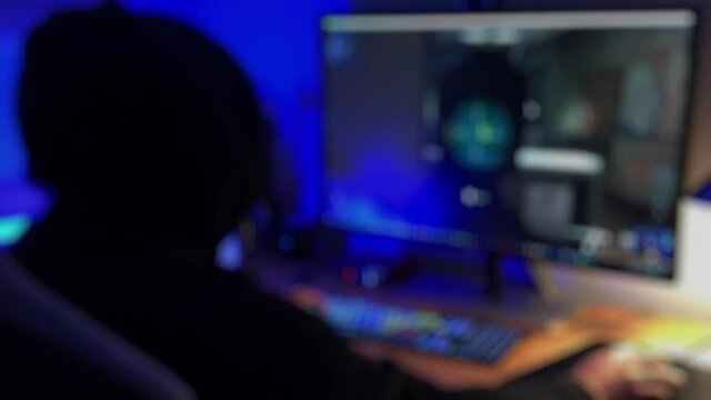 Teenage PC Gamer Playing First-Person Shooter in Subdued Light. A teenage gamer dressed in a black hoodie plays a multiplayer first-person violent shooter video game on a PC.