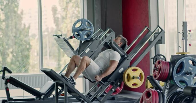 Sporty man doing exercises with leg press machine at fitness gym, hard squats workout