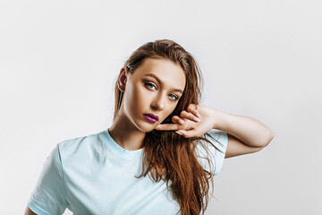Beautiful young brunette girl with green eyes with long hair with fashion makeup with purple lips on an isolated gray background. The woman holds hand to face and seriously looking at the camera.