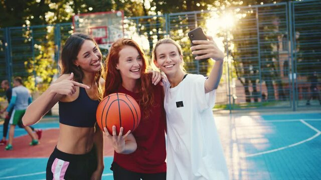 Sports team. Girl power. Athletic beautiful three girls taking picture selfies on smartphone holding basketball ball training in sportground. Active people. Females. Social media.