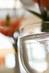 a vertically oriented closeup shot of a glass of water in the foreground with flowers blurred in the background in a restaurant one bright sunny morning 