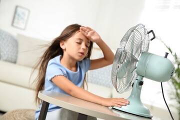 Little girl suffering from heat in front of fan at home. Summer season