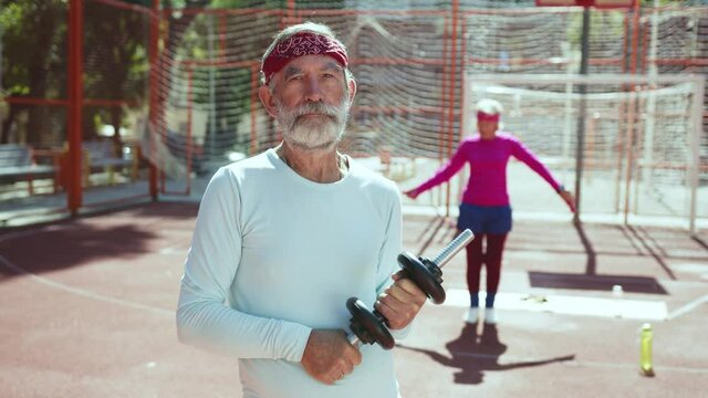 Old active athletic man with dumbbells posing for camera portrait standing in basketball court. Ederly woman working out in background. Families in sports.