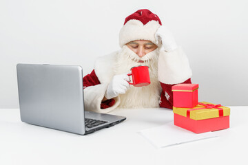 Santa Claus is sitting at his desk, drinking coffee and working at the computer. There are Christmas gifts on the table.