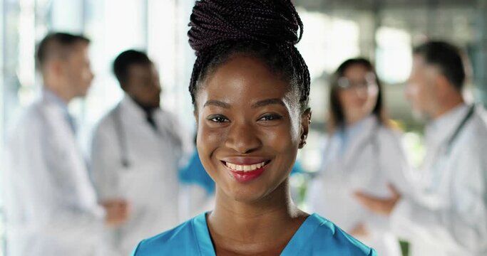 Close up of African American happy beautiful woman physician smiling to camera in clinic. Inside. Portrait of cheerful pretty female doctor or nurse in hospital. Mixed-races docs talking on background
