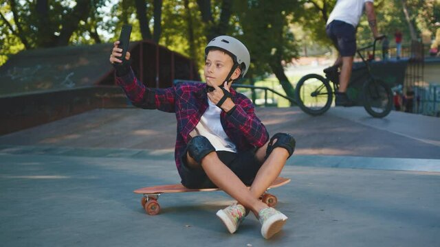Teenager happy skater boy in helmet sitting on a skate and taking funny selfie picture. Modern teen kid taking photographs on smartphone in skatepark.