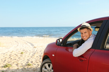 Happy man leaning out of car window on beach, space for text. Summer trip