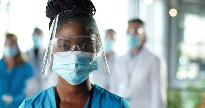 Close Up Of African American Woman Physician In Face Shield Looking To Camera In Clinic. Portrait Of Female Doctor Or Nurse In Medical Mask And Goggles In Hospital. Docs Talking On Background Covid-19