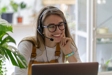 Smiling gardener millennial woman wear headphones talk video calling, using laptop, communicating...