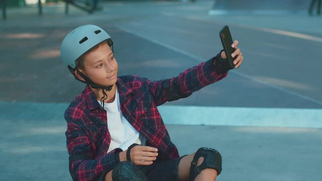 Happy teenager taking selfie picture smiling on camera sitting in the skatepark. Skateboarder boy. Smartphone communication. Social media. Outdoor activities.