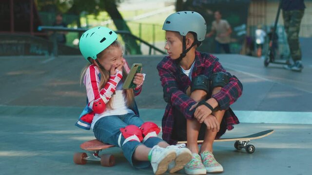 Funny girl playing with brother in skatepark taking funny face pictures on smartphone. Siblings having fun. Excited girl sitting riding on skateboard.