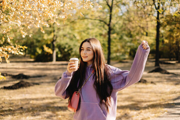 Beautiful young woman having fun, drinking coffee and enjoying autumn weather in the park. Woman walking in the autumn park.