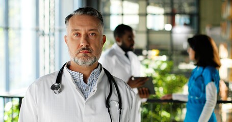 Portrait of Caucasian serious man physician in white gown looking at camera and standing in clinic. Indoors. Handsome male doctor in hospital. Mixed-races doctors talking on background.