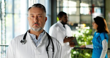 Portrait of Caucasian serious man physician in white gown looking at camera and standing in clinic. Indoors. Handsome male doctor in hospital. Mixed-races doctors talking on background.
