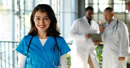 Portrait of Caucasian happy beautiful woman physician smiling to camera and holding documents in clinic. Indoor. Pretty female doctor in hospital. Multi ethnic doctors talking on background.