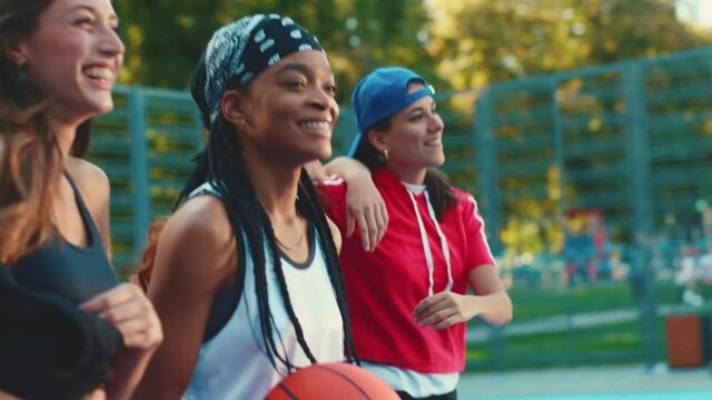 Multi-ethnic team of attractive girls walking inside basketball court bouncing ball chatting smiling having fun sportive games together. Female players. Active lifestyle.