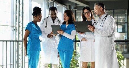 Multi ethnic doctors, men and women, standing in hospital, talking and discussing diagnosis. Mixed-races males and females physicians having discussion with folder of documents and tablet device.