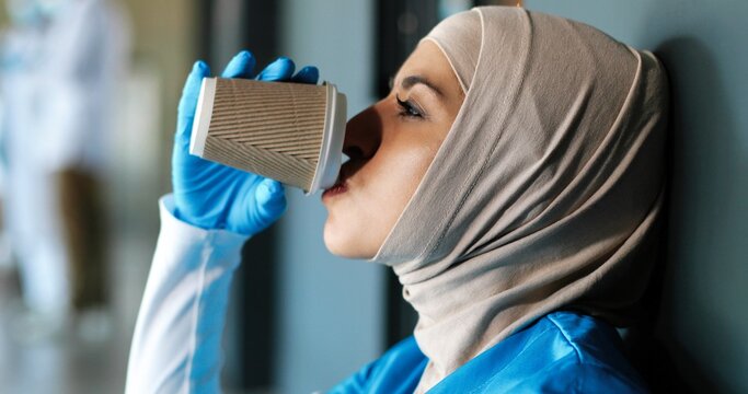 Close up of tired Arab young woman doctor in hijab and gloves drinking coffee and leaning on wall. Muslim female nurse in headscarf resting, having break, sipping drink. Coronavirus work. Having rest.