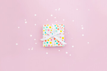 Gift box decorated with white bow and paper with bright colored polka dots on pink background with star confetti. Selective focus