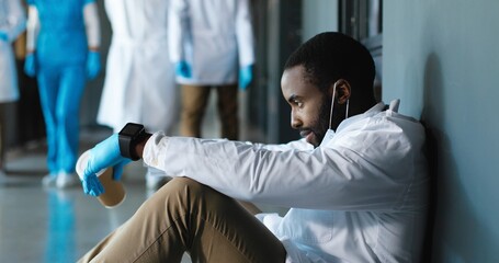 Tired sad African American man doctor in medical mask and gloves drinking coffee and leaning on...