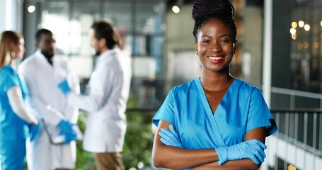 Portrait of African American happy beautiful woman physician in gloves smiling to camera in clinic. Joyful pretty female doctor or nurse in hospital. Mixed-races docs talking on background.