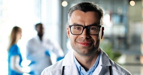 Portrait of Caucasian man physician in white gown and glasses looking at camera, smiling and standing in clinic. Handsome male doctor in hospital. Doctors on background. Medicine concept.