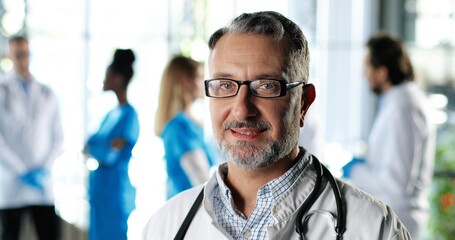 Fototapeta premium Portrait of Caucasian senior man physician in white gown, stethoscope and glasses looking at camera and smiling in clinic. Handsome male doctor in hospital. Doctors on background. Healthcare.