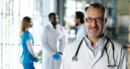 Portrait shot of Caucasian senior man physician in white gown, stethoscope and glasses looking at camera and smiling in clinic. Handsome male doctor in hospital. Doctors on background Medicine concept