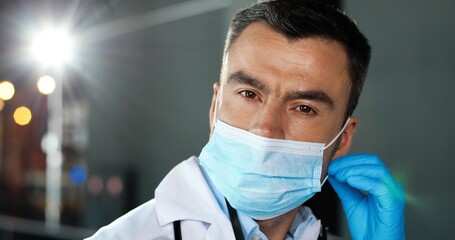 Close up of Caucasian man physician taking off medical mask and goggles looking at camera in clinic. Portrait of male doctor in hospital. Docs talking on background. Covid-19 concept. Pandemic.