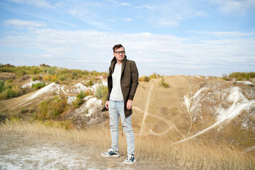 Serious man with glasses in jeans, t-shirt and cape stands in hilly terrain. Adult male with eyeglasses in hood looks away thoughtfully in countryside.