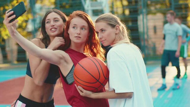 Trio of beautiful slim cheerful girls taking group selfie picture on smartphone training playing basketball game on court. Sports team. Sexy women. Best friends.
