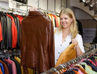 Positive girl looking for new leather jacket during shopping in retail shop