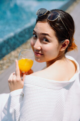 Asian woman traveler in white bathrobe enjoy drinking orange juice at poolside outdoors.