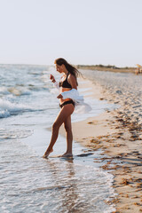 Fototapeta na wymiar Young beautiful slender woman in a black bikini and a white shirt on a tanned body is walking on the beach. Soft selective focus, art nose.