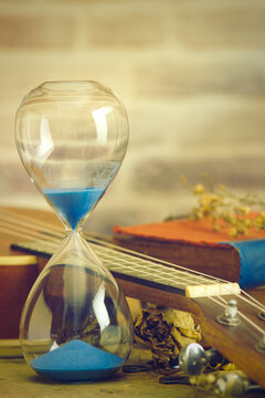 A vintage hourglass and ukulele with an old book and brass pen on a wooden table and brick background in the morning. Closeup and copy space. The concept of memories or things in the past.