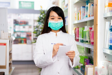 Diligent friendly smiling chinese woman pharmacist in protective facial mask keeps track of drugs in interior of pharmacy