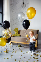 Cute blonde boy on his first birthday at a decorated party in gold, black and silver colors. Playing with balloons.