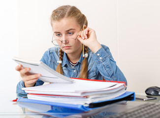 Naklejka premium Concentrated young girl doing her homework with laptop and notebook at kitchen table