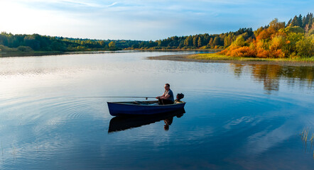  Elderly man fishing with a rod on a small fishing boat on the lake at autumn sunset - Powered by Adobe