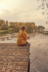 A girl in a yellow knitted sweater sits on a wooden bridge over the lake and catches fish.