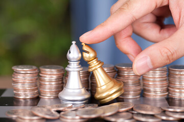 A hand in front of a large number of dollar coins helps the silver chess piece to defeat the golden...