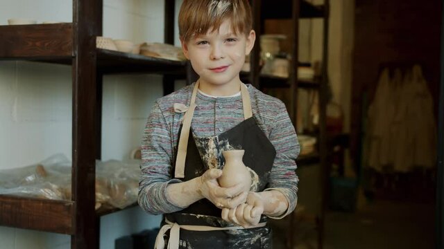 Portrait of joyful boy young potter holding hand-made vase in workshop standing alone smiling looking at camera. Child is wearing dirty apron.