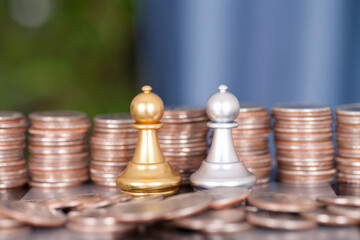 In front of a large number of dollar coins, one hand helps the silver chess piece to defeat the golden chess piece