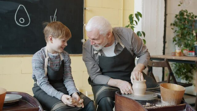 Grandfather potter is making vase on throwing-wheel and talking to cheerful kid in studio enjoying creative hobby and communication with grandchild.