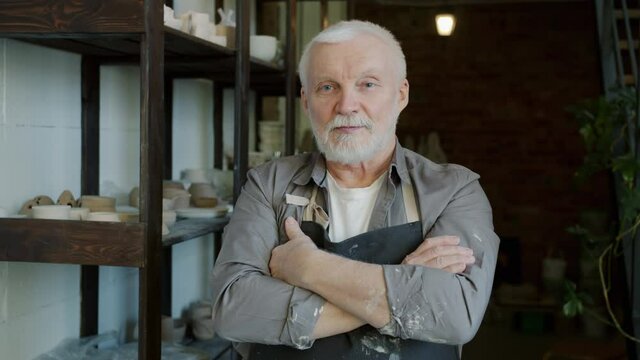 Portrait of handsome elderly man potter standing in workshop wearing apron and looking at camera with shelf with hand-made ceramics in background