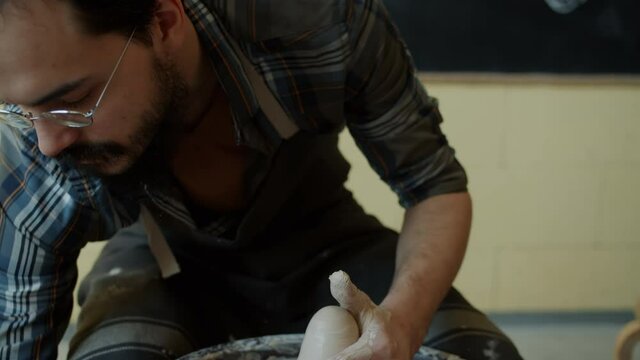 Skillful young potter is molding ceramic vase from clay on turning throwing wheel working in studio. Professional is concentrated on creative work.