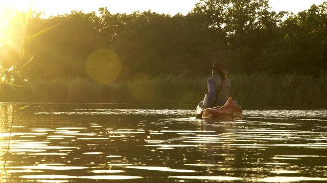 A cowboy in a red canoe floats on the river in the forest. Beautiful sunlight at sunset. Historical reconstruction of life in the wild west of America. 4K